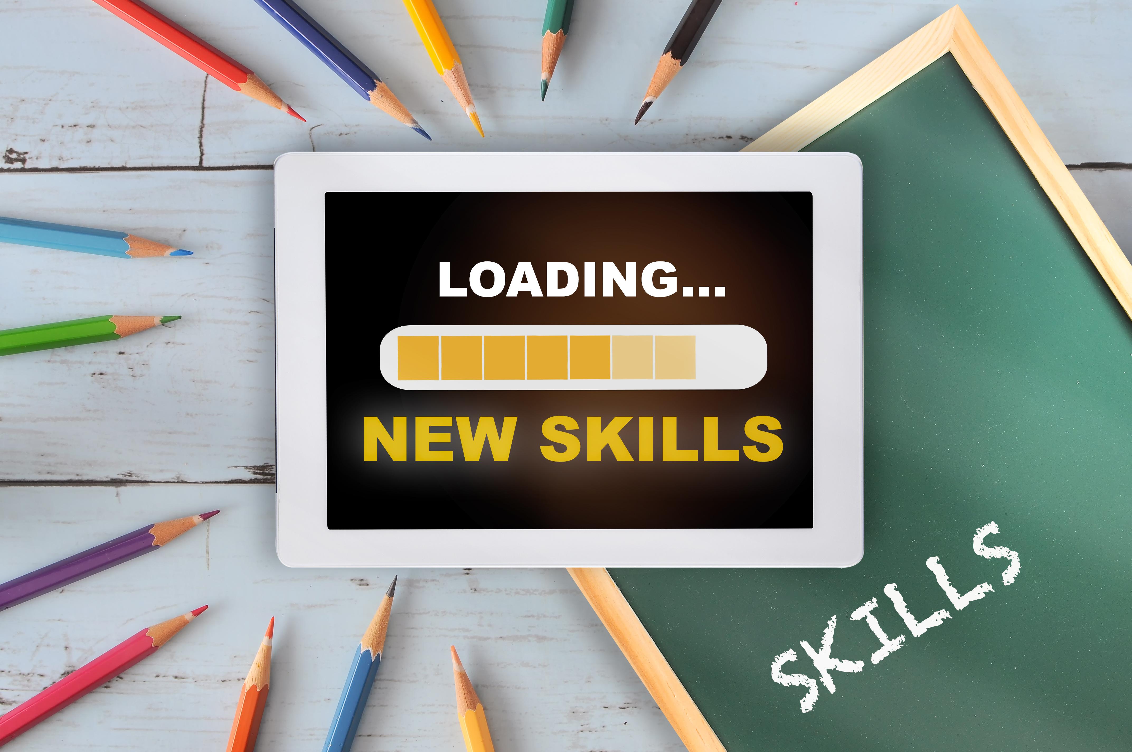 A tablet with the words ' loading... new skills' is on a background of coloured pencils and a green chalkboard.