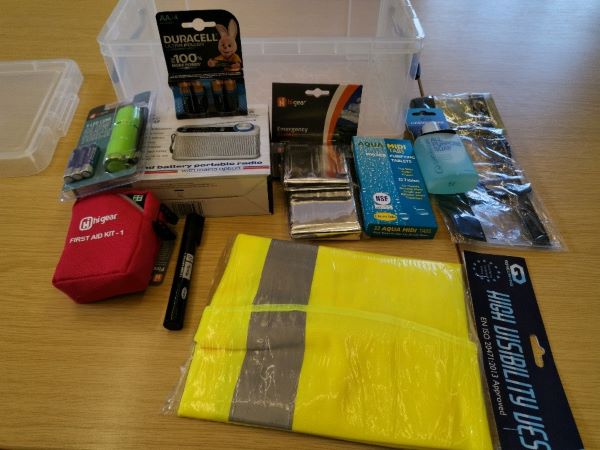 A photo of a plastic box, flashlight, a battery-powered radio and batteries, hi visibility jacket, emergency blanket, water-purifying tablets, soap and a first aid kit.